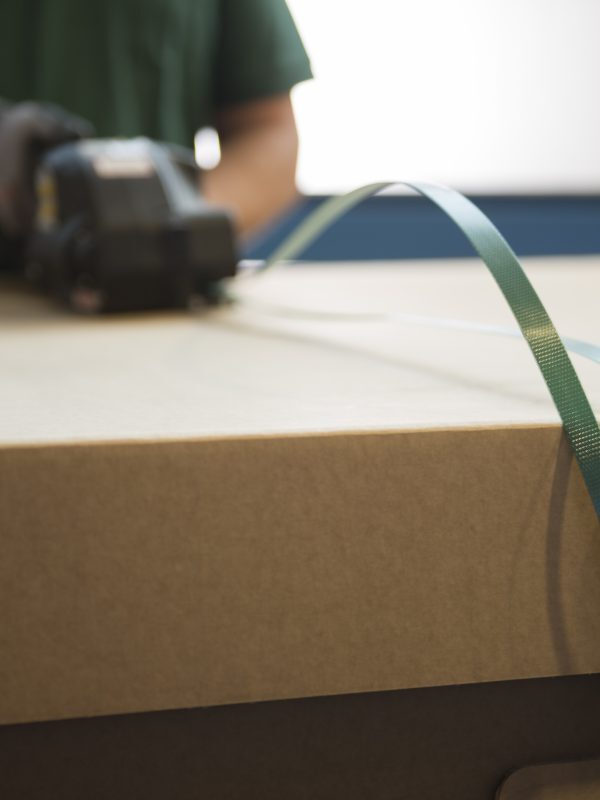 Mitarbeiter spannt Umreifungsbänder um eine großformatige Kartonverpackung